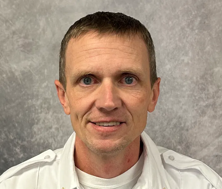 A person smiling against a gray textured background, wearing a white shirt.