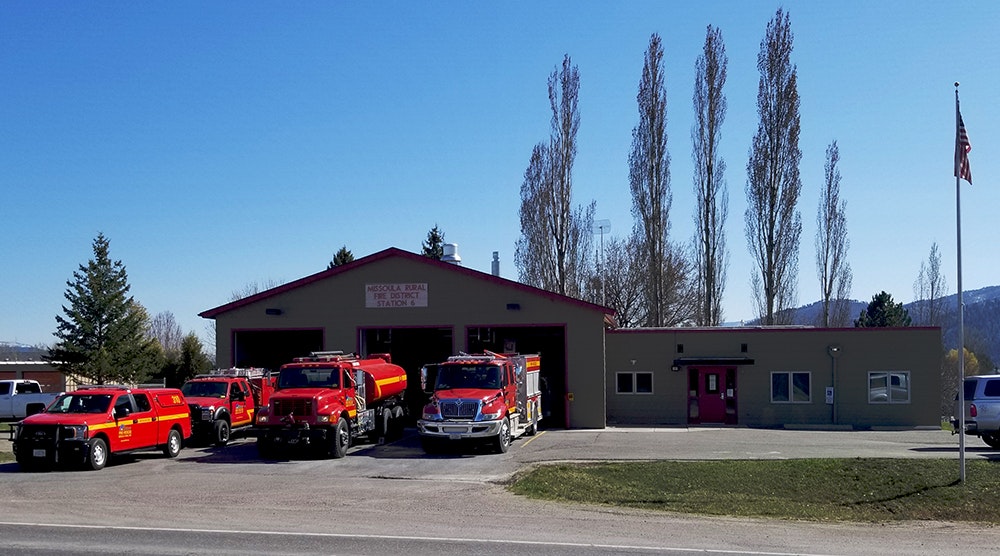 A fire station with several red fire trucks parked outside, surrounded by trees and a flagpole.