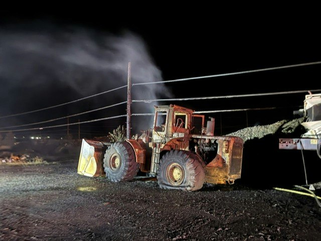 An old, rusty bulldozer emits steam at night, with power lines and a pile of material nearby.