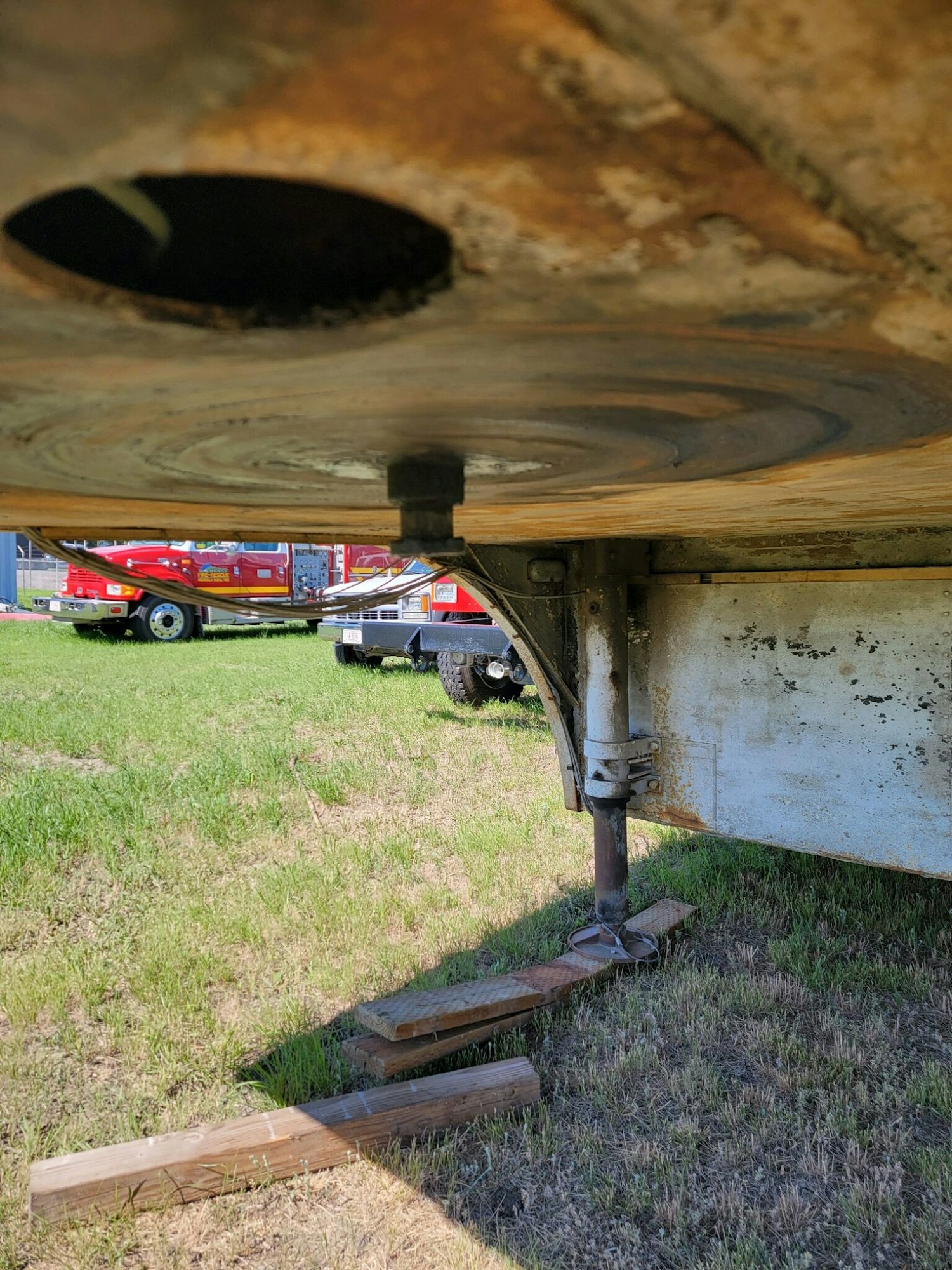 Underside of a trailer's hitch area with a landing gear leg, supported by wooden blocks, grass, and a red vehicle in the background.
