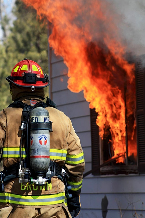 A firefighter in gear stands near a house window with flames pouring out.