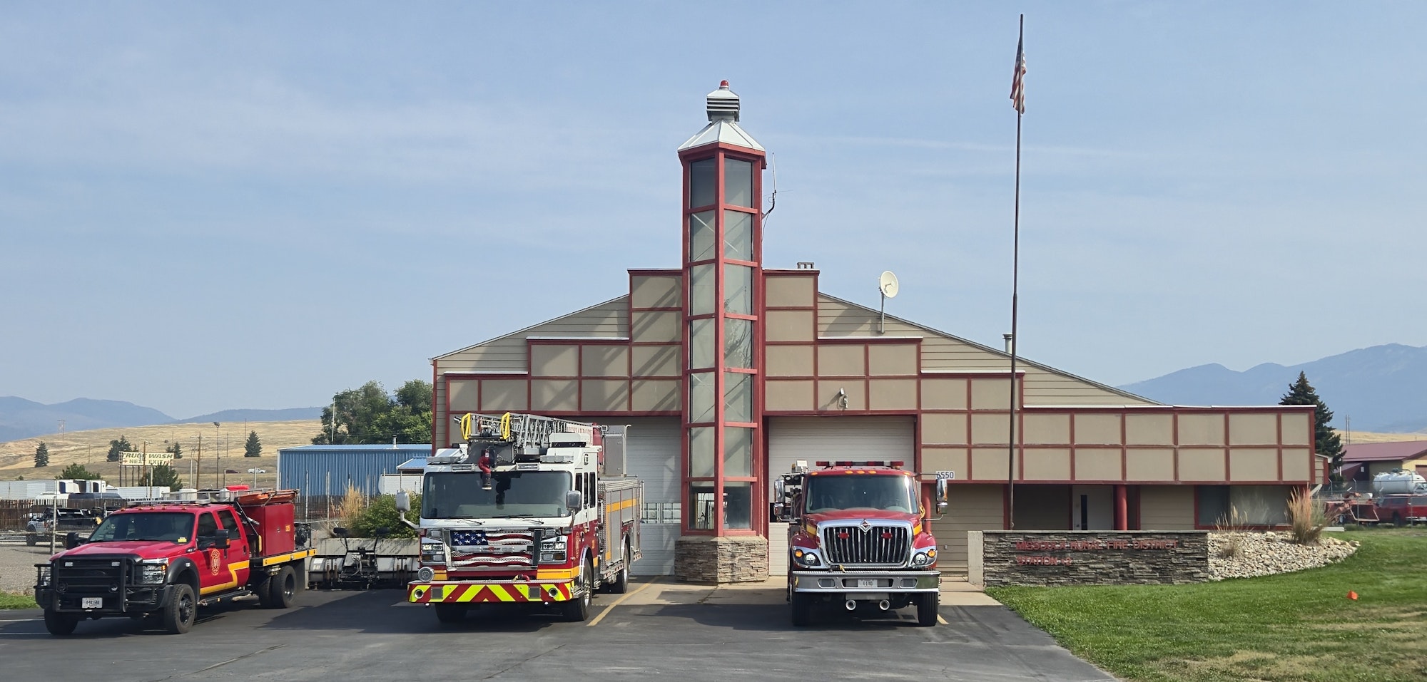 The image shows a fire station with two fire trucks parked in front, set against a backdrop of mountains and clear skies.