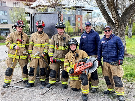 A group of firefighters stands together outdoors, wearing uniforms and holding equipment.