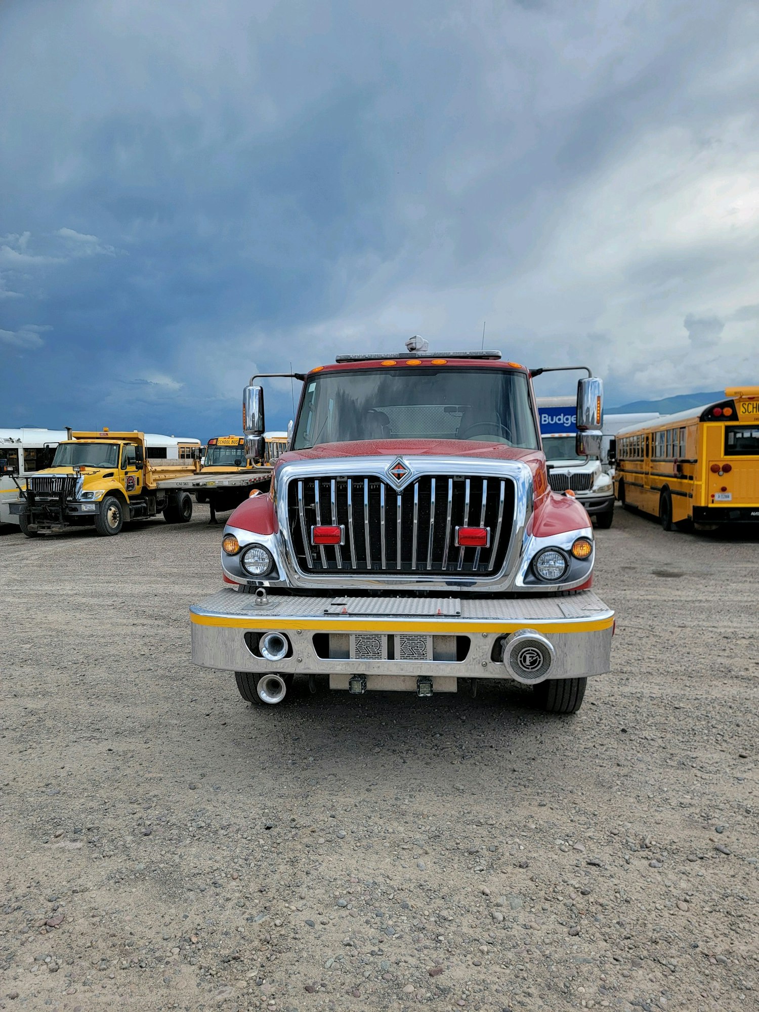 A red fire truck parked on gravel with school buses in the background under a cloudy sky.