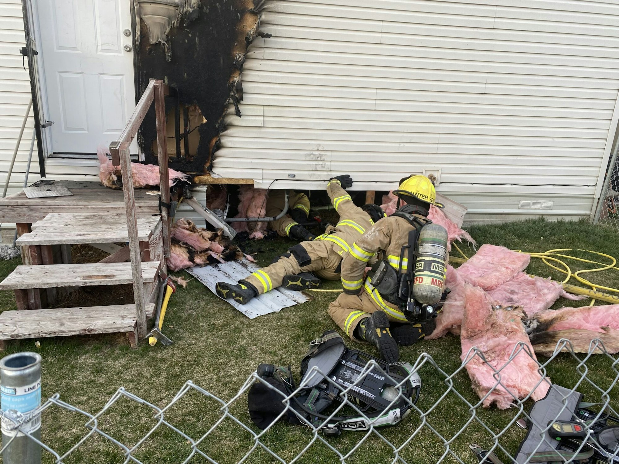 Firefighters examining a burned area on the exterior of a building, with insulation removed and firefighting equipment nearby.