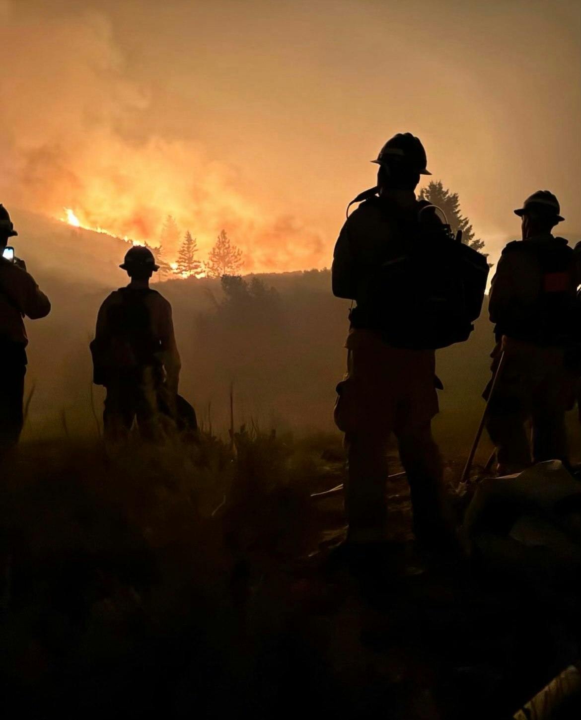 Firefighters silhouetted against a large wildfire at night.