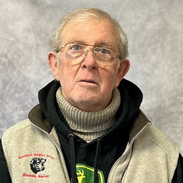 A person wearing glasses, a gray fleece vest, and a black hoodie with a gray turtleneck underneath, against a soft gray background.