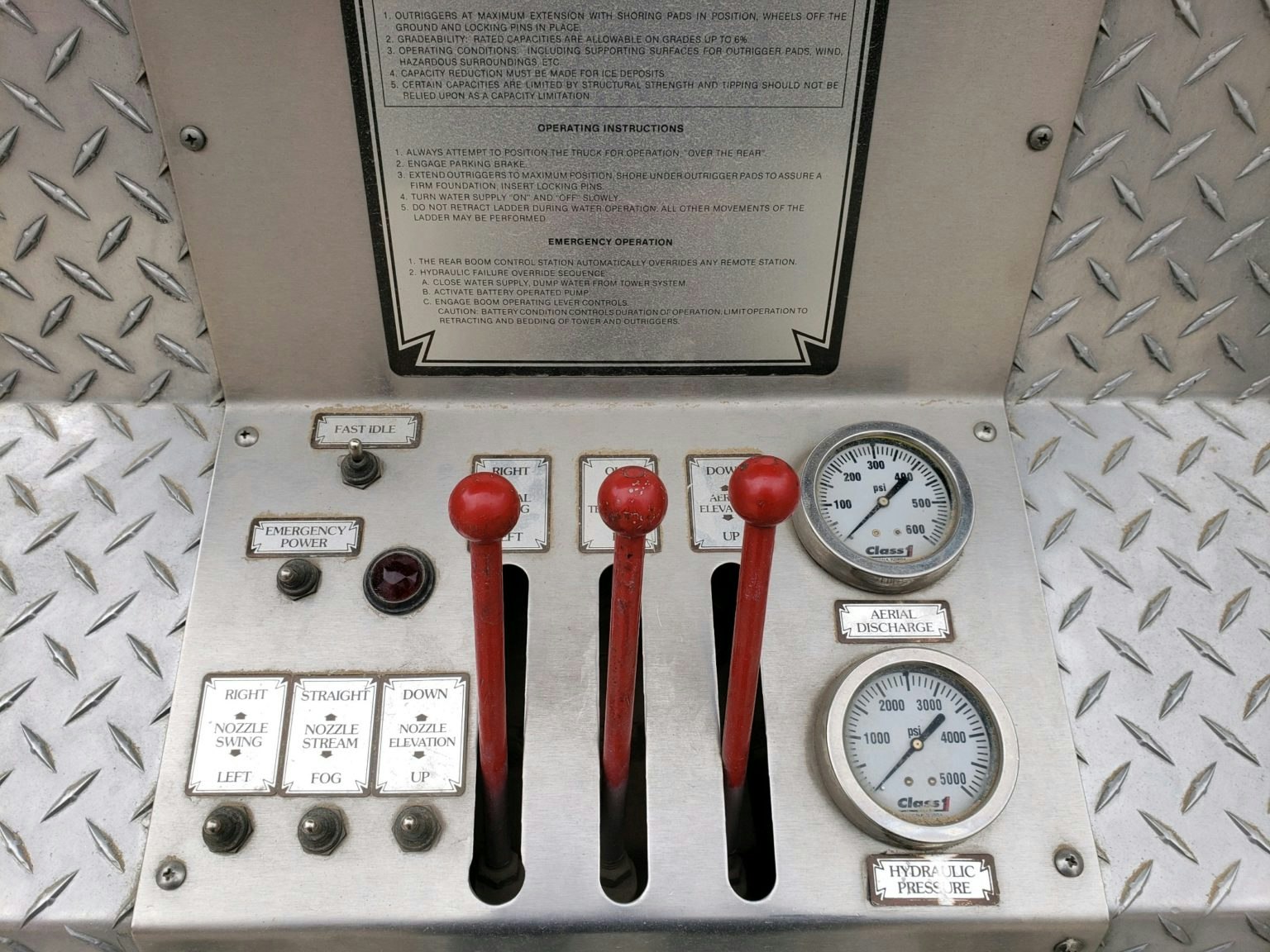Control panel with levers, gauges, and switches for operating a fire truck or equipment, set against a diamond plate background.