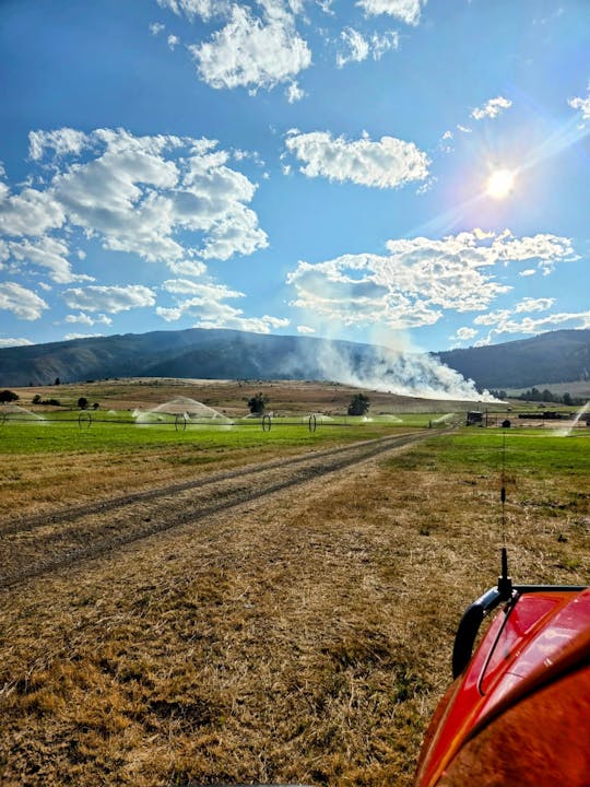 A sunny landscape with clouds, hills, sprinklers, and a red vehicle on dry grass. Smoke is visible in the distance.