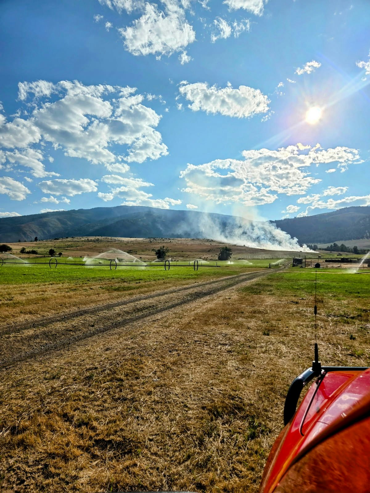 A sunny landscape with clouds, hills, sprinklers, and a red vehicle on dry grass. Smoke is visible in the distance.