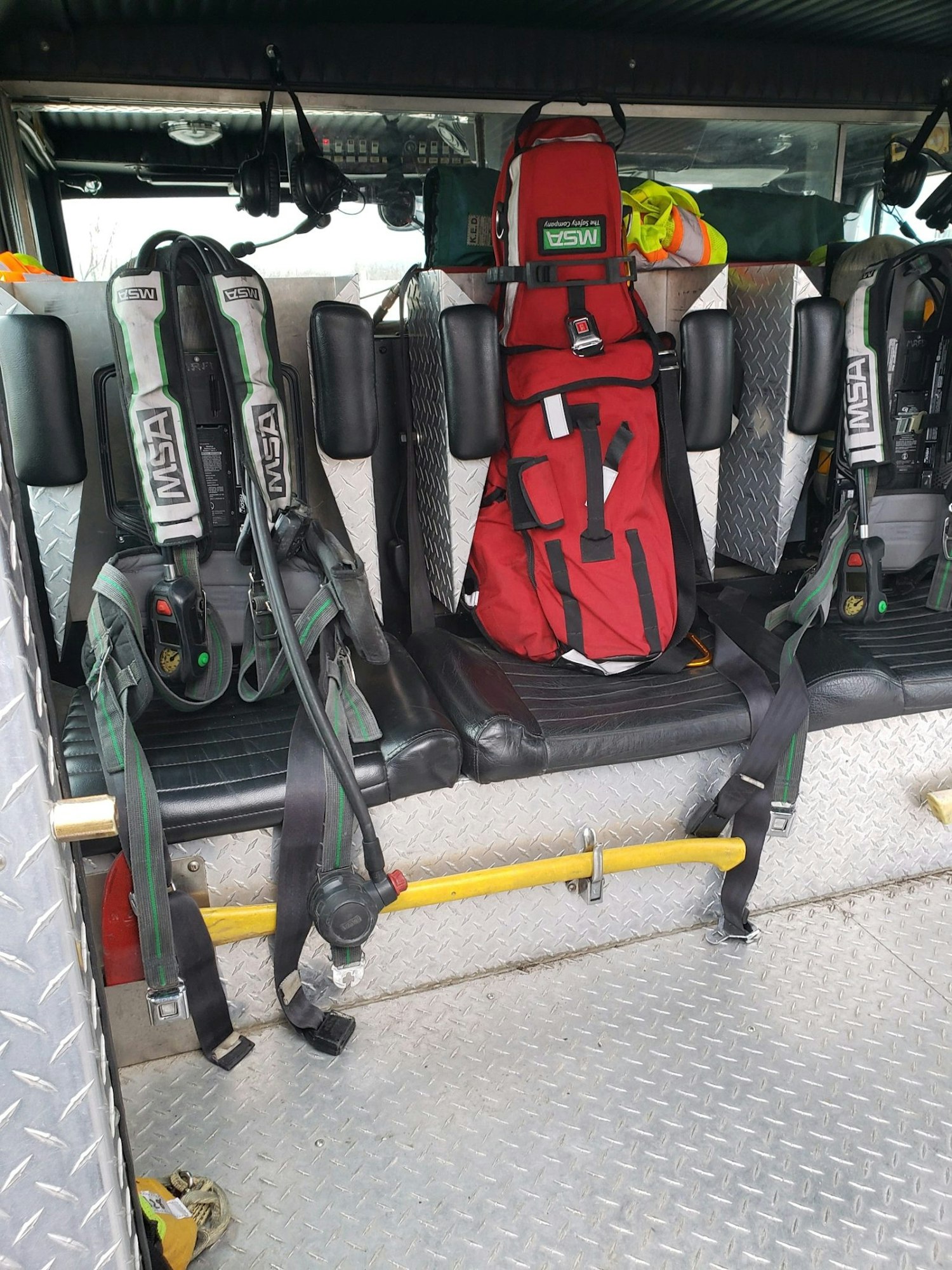 Firetruck interior with MSA breathing apparatus, red emergency bag, yellow safety bar, and seating on diamond plate floor.