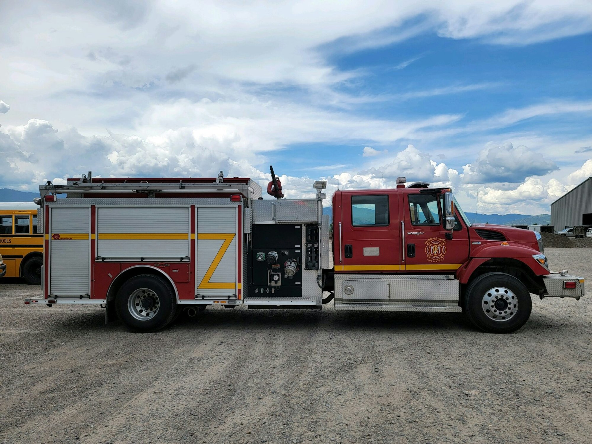 A red fire truck parked on a gravel surface with a cloudy sky and a school bus in the background.