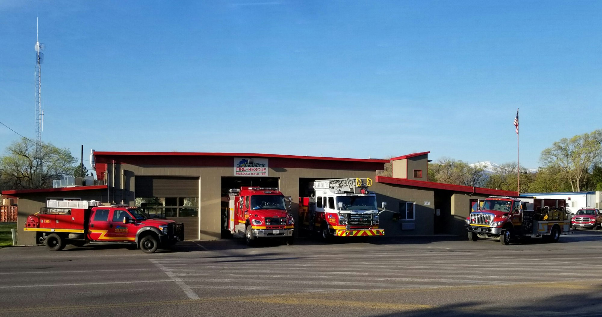 Fire station with three fire trucks parked outside, American flag, and clear blue sky.