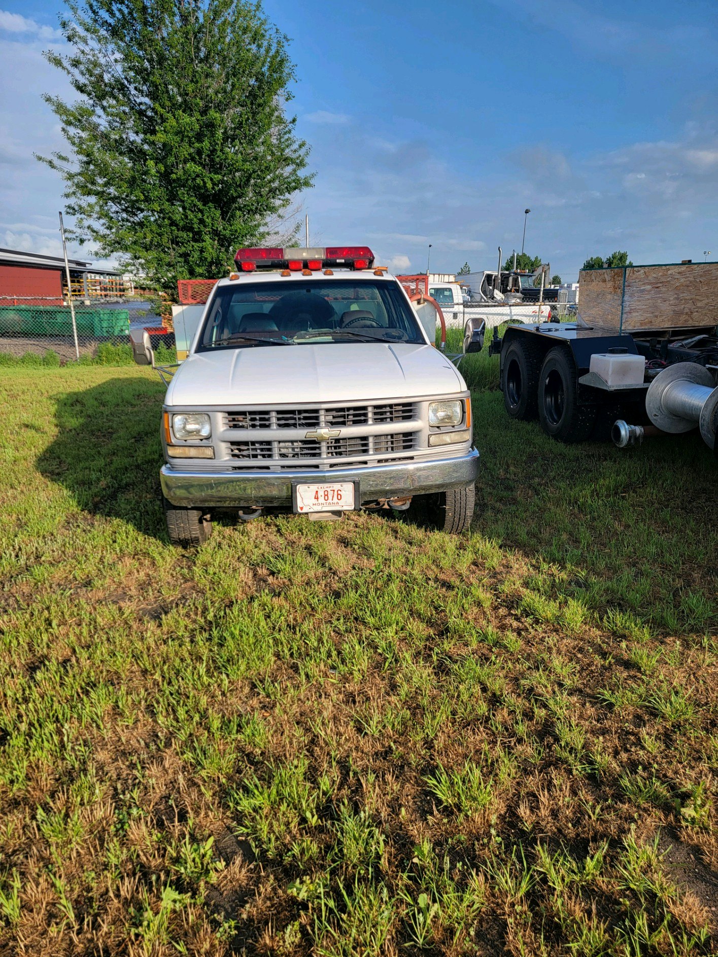 A white utility truck with red lights parked on grass next to a larger vehicle and a tree in the background.