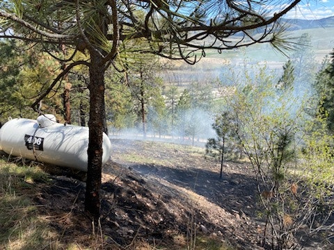 A propane tank near a scorched hillside with smoke in a forested area.