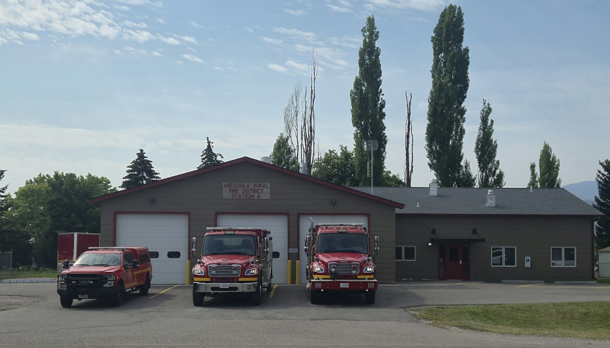 Image shows Missoula Rural Fire District Station 6, featuring two fire trucks, a building, and trees in the background.