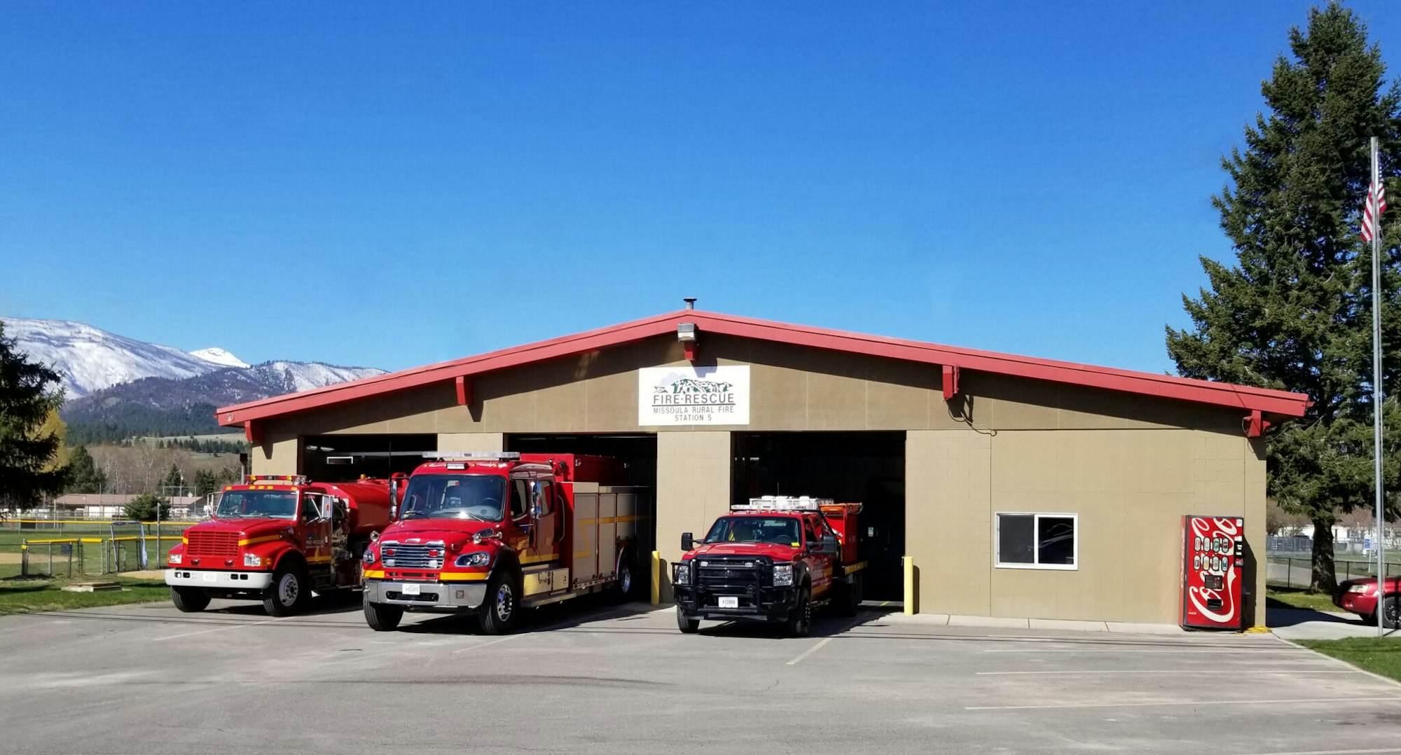 Fire station with fire trucks in front, set against a mountainous background.