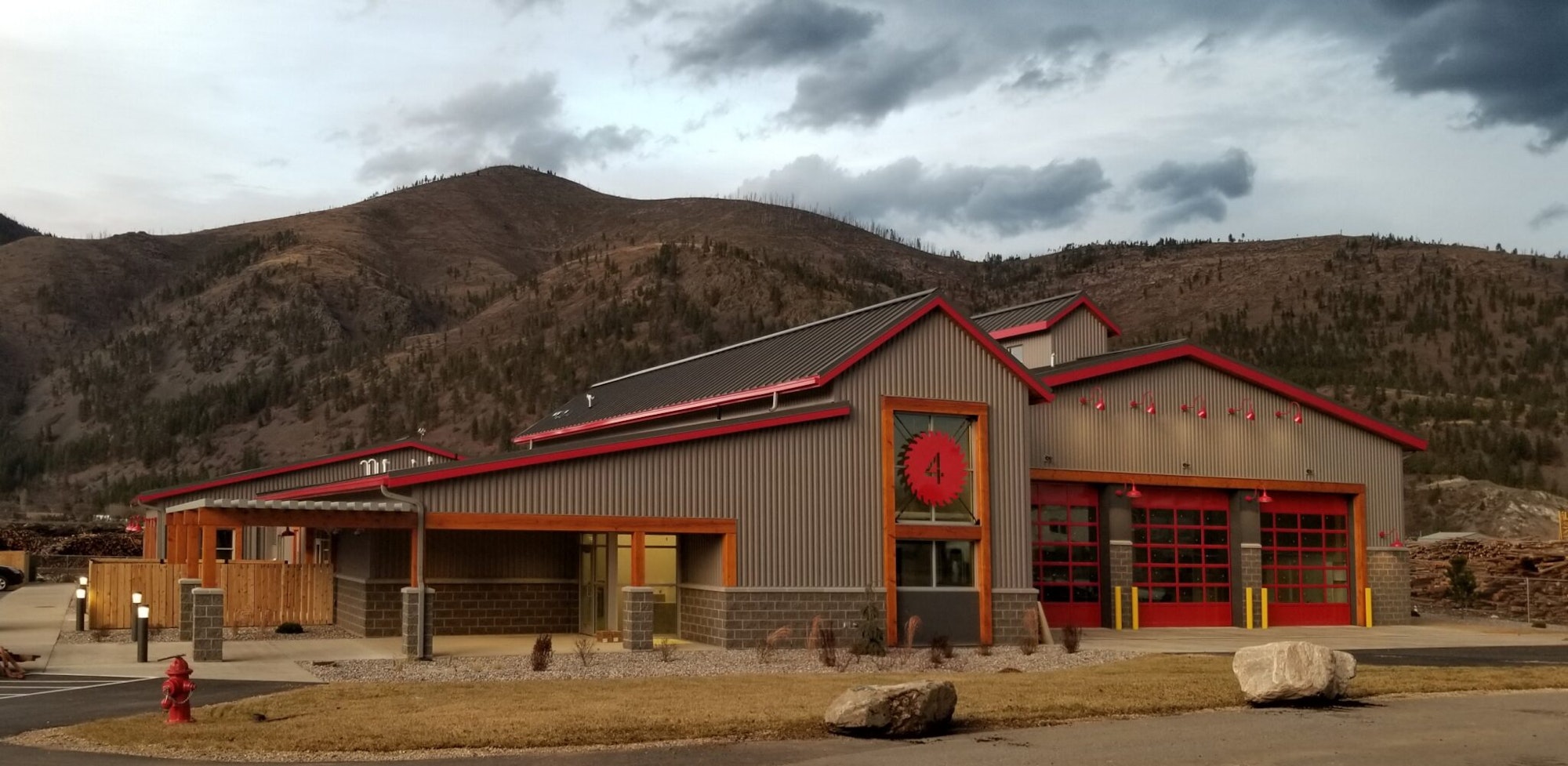 A modern fire station with a red and gray exterior, surrounded by hills and cloudy skies.