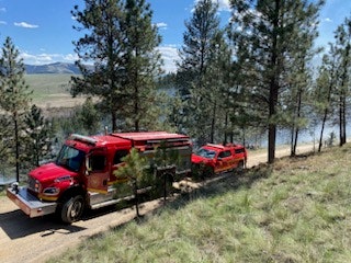 Two red fire vehicles on a grassy riverside path surrounded by trees under a blue sky.