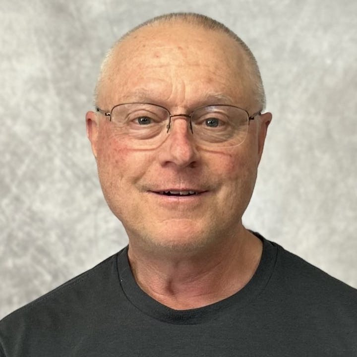 A man with glasses and a black shirt, smiling against a gray background.