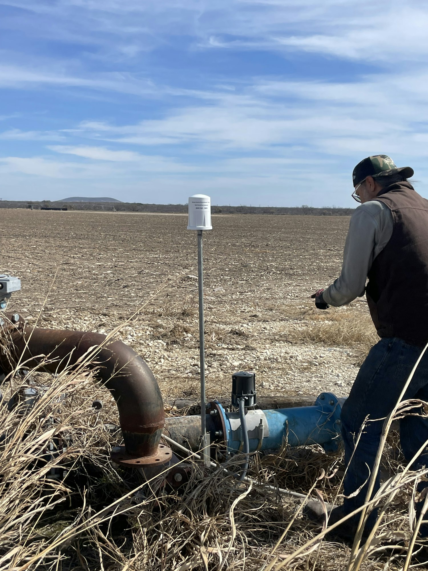 An individual is near a water pump in a field, measuring or inspecting something, with a clear sky in the background.