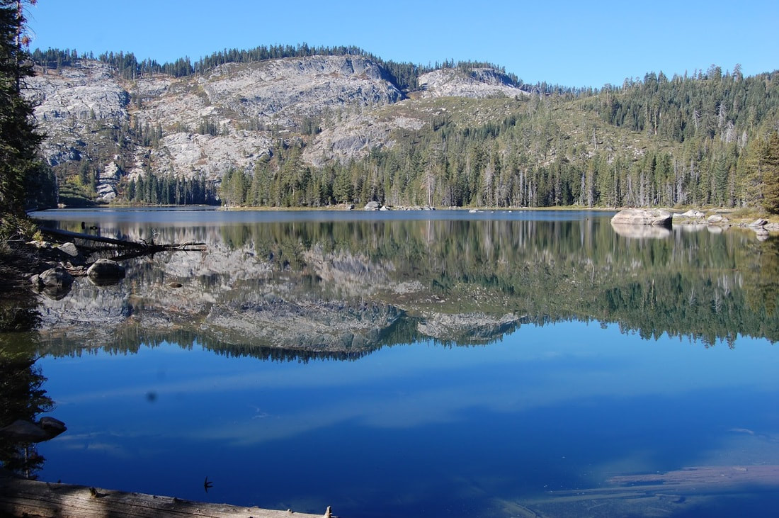 A serene mountain lake reflecting surrounding trees and rocky hills, under a clear blue sky.