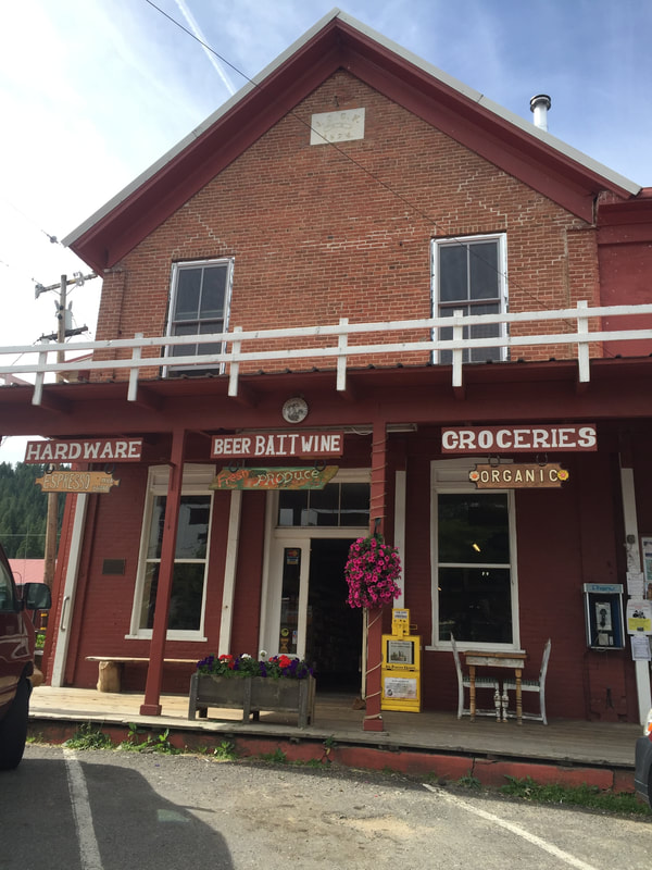 A rustic red brick building with signs for hardware, groceries, and beer. It features a porch with flowers and chairs.