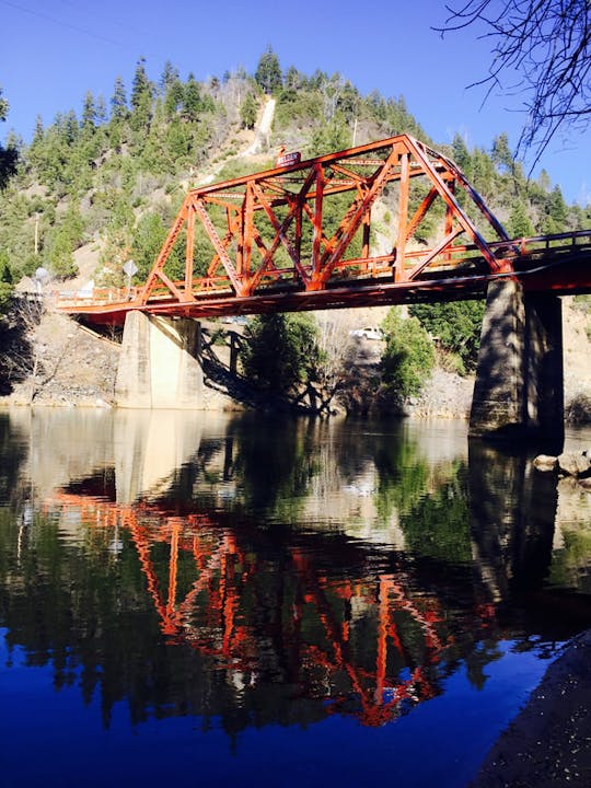 A red truss bridge over a calm river with its reflection visible, surrounded by trees.