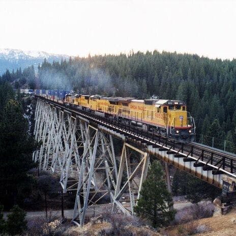 A freight train crosses a steel bridge in a forested, mountainous area.