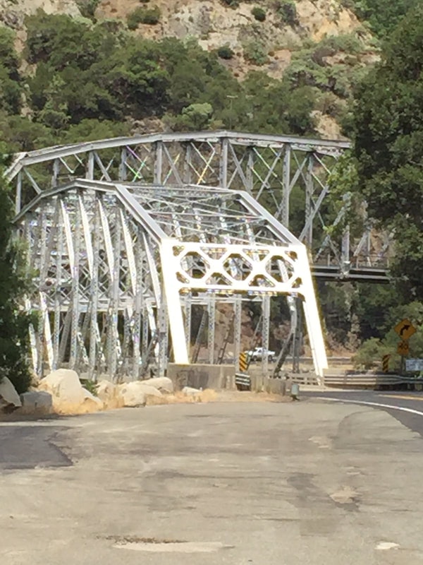 A metal truss bridge with a road leading to it, surrounded by rocky hills and trees.
