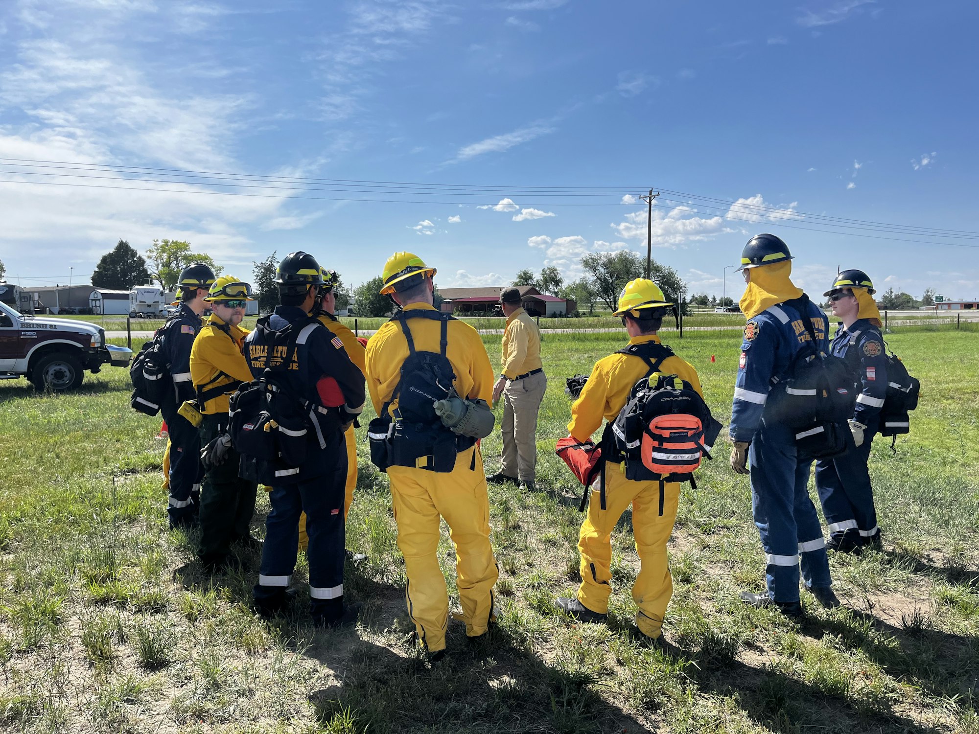 A group of firefighters in yellow and blue uniforms, standing on grass, receiving instructions near a fire truck.