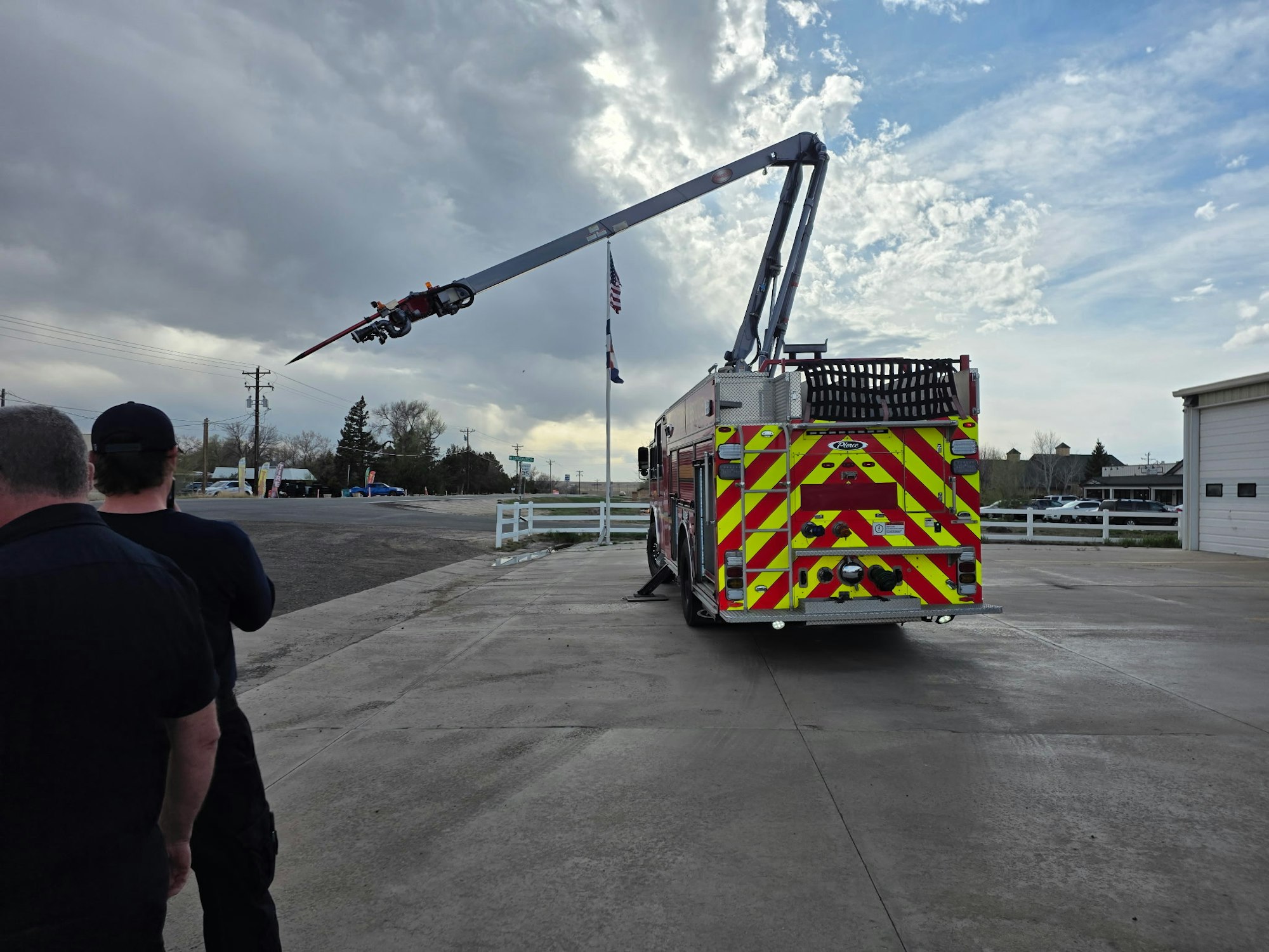 A fire truck is seen with its ladder raised, while two people watch from behind in a mostly cloudy outdoor setting.