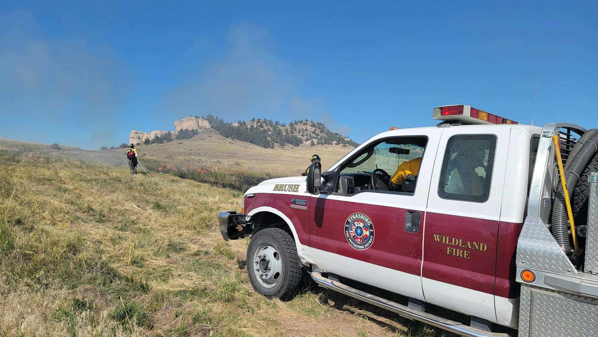 Firefighters in a field with a "Wildland Fire" truck, smoke in the background, and rocky hills in the distance.