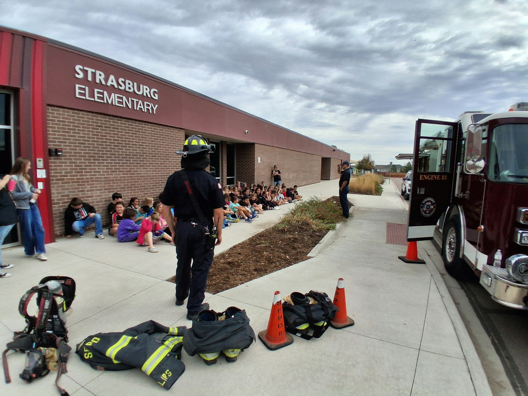Firefighters and a fire truck outside Strasburg Elementary, with students gathered on the sidewalk for a demonstration or drill.