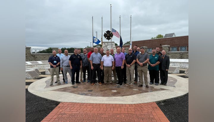 A group of people stands in front of a monument with flags, plaques, and a brick pathway in an outdoor setting.