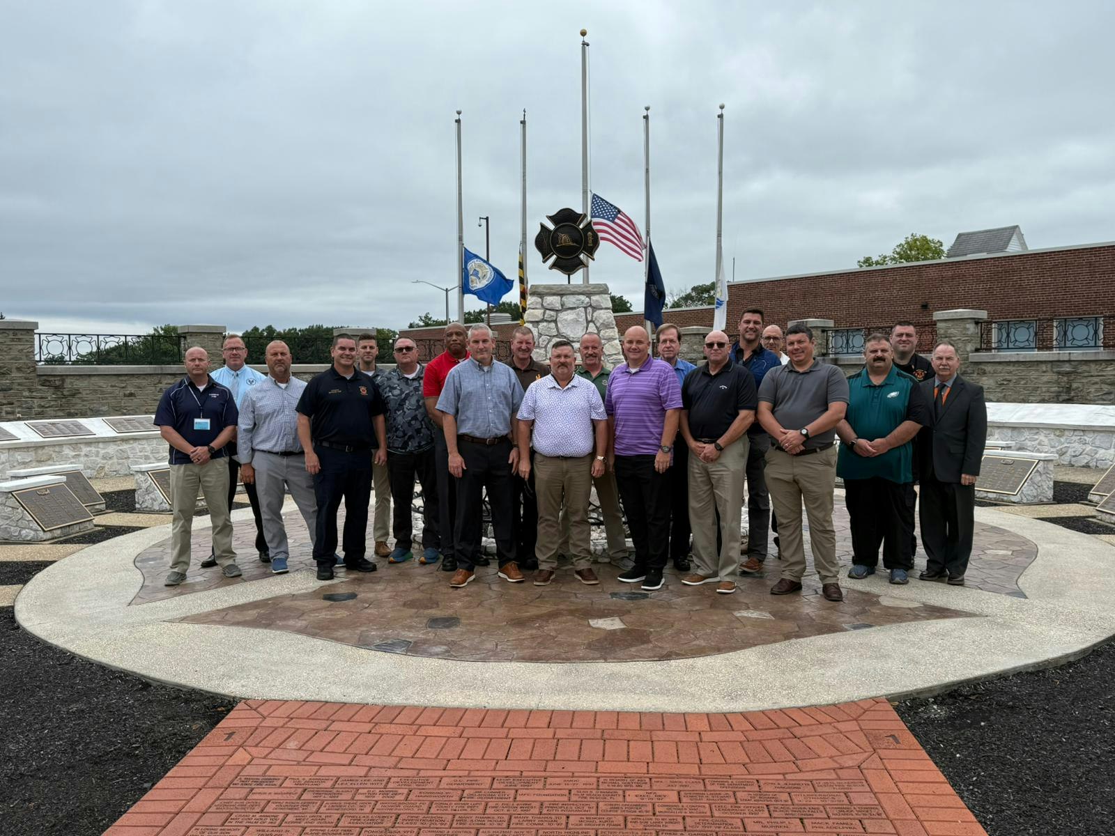 A group of people stands in front of a monument with flags, plaques, and a brick pathway in an outdoor setting.
