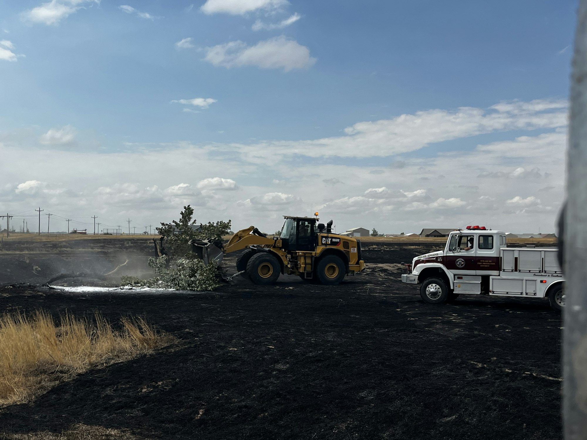 A bulldozer moves trees near a fire truck on a scorched field under a partly cloudy sky.