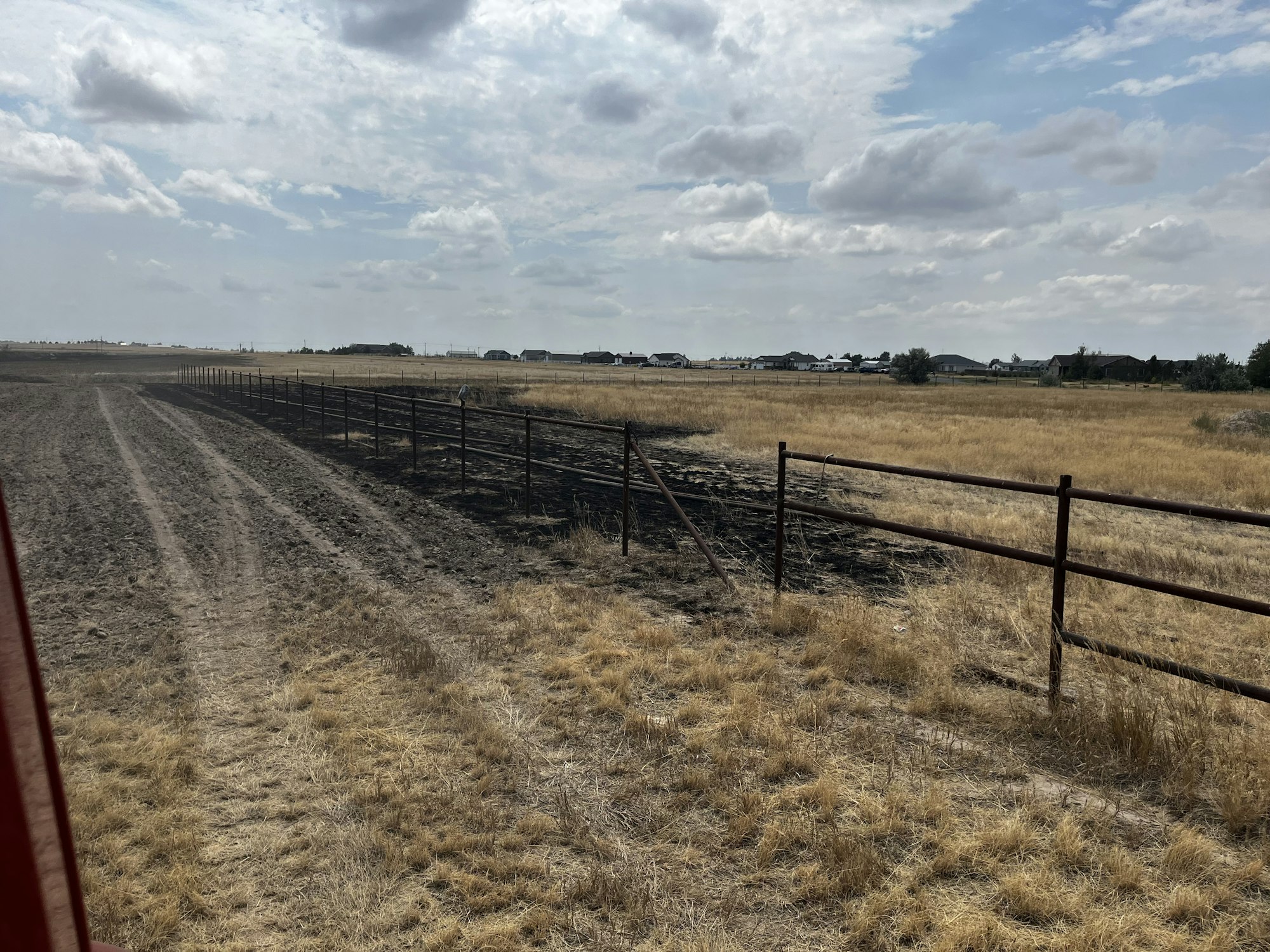 A dry field with a metal fence, dirt road, and cloudy sky, surrounded by houses in the distance.