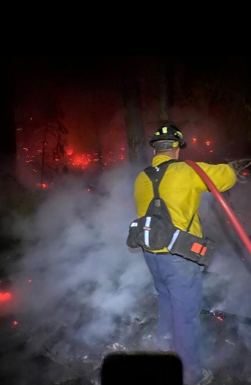 A firefighter amidst smoke, facing a forest fire at night, holding a hose.