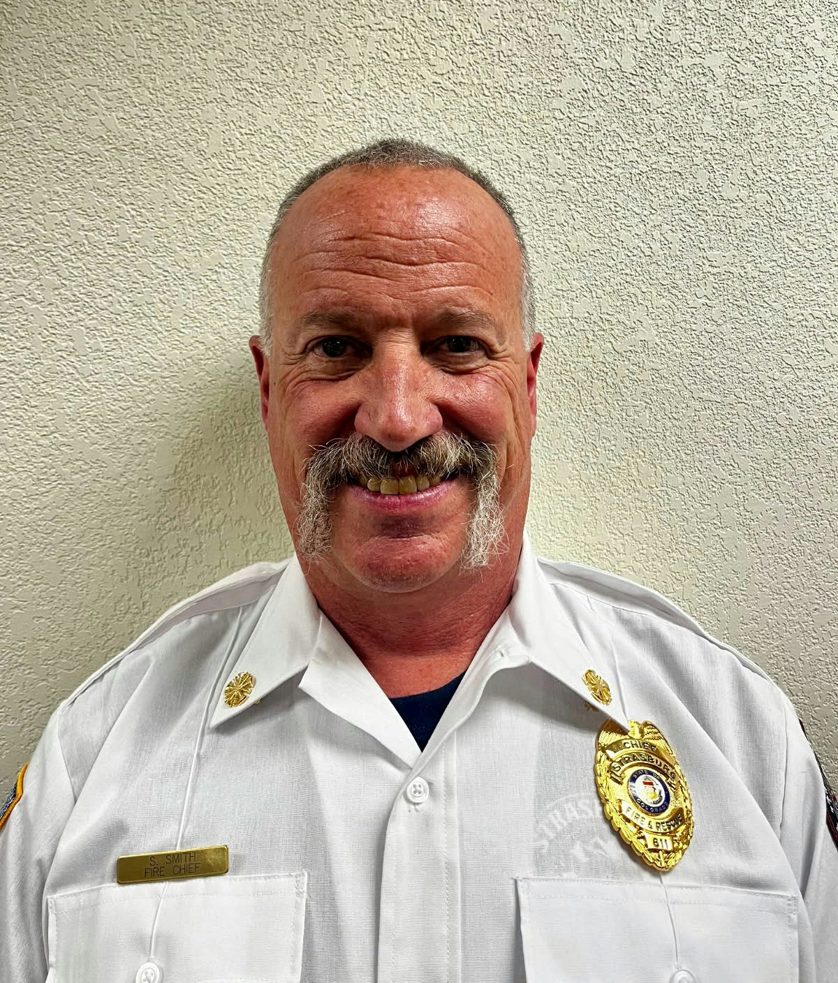 A person in a white uniform with badges and a nametag, smiling at the camera.