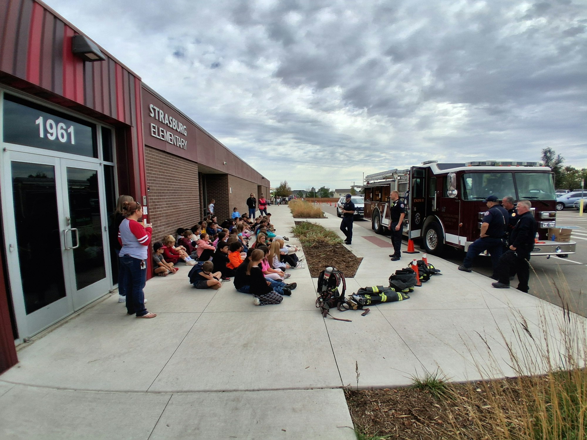A group of children sitting outside Strasburg Elementary near a fire truck and firefighters.