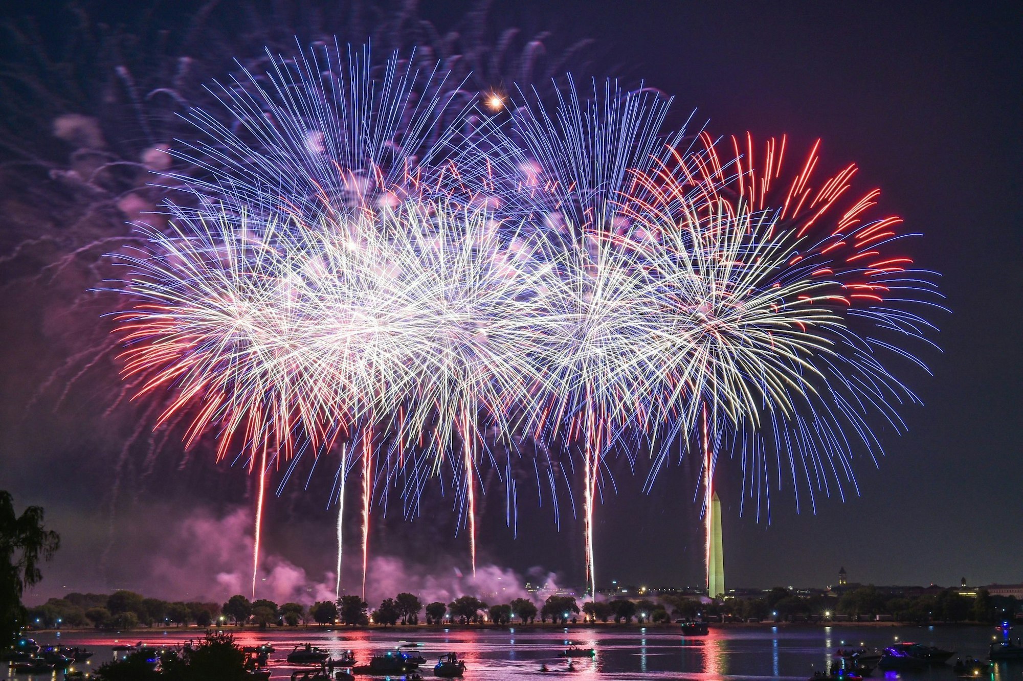 Colorful fireworks exploding over a river at night with silhouettes of boats and trees in the foreground.