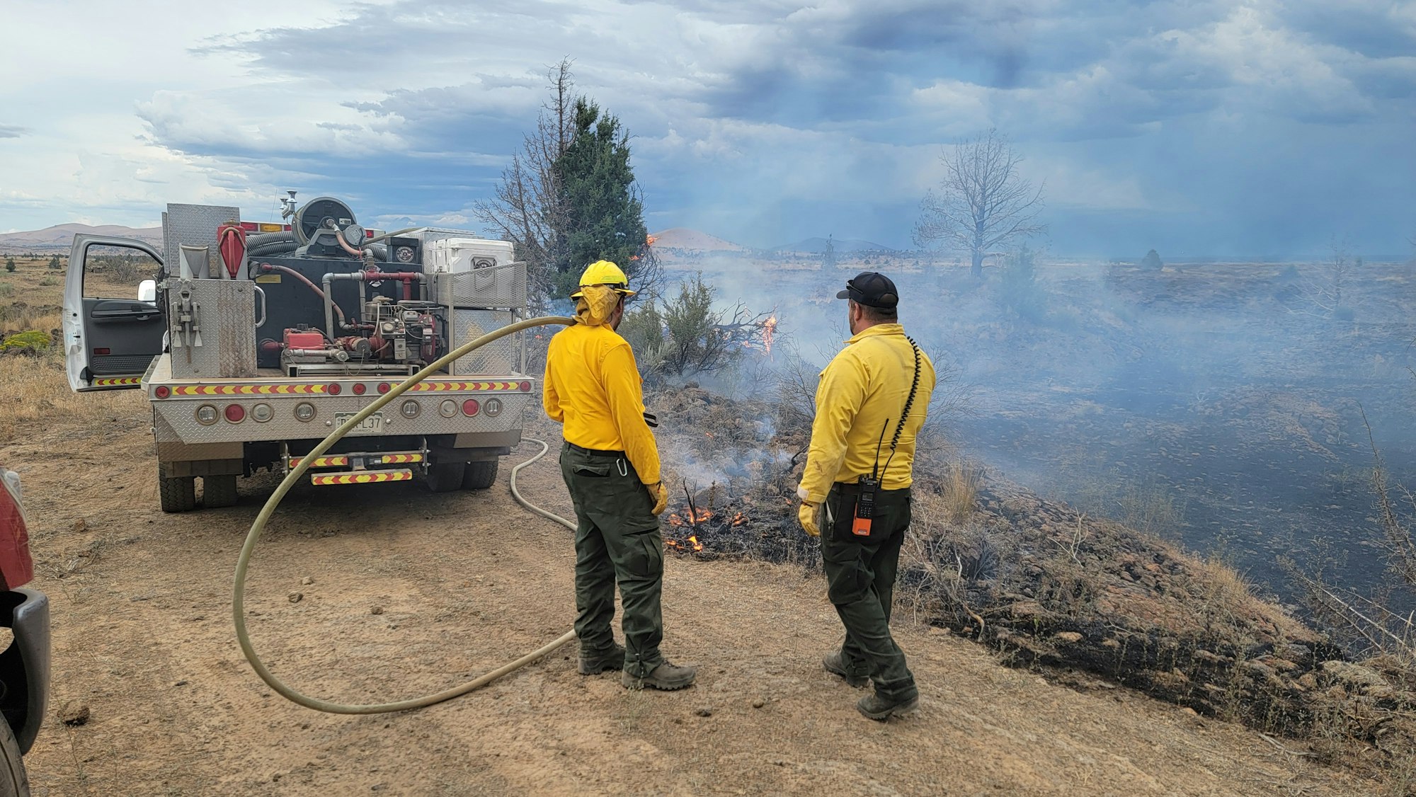 Two firefighters in yellow shirts stand near a fire truck, managing a brush fire with smoke rising.