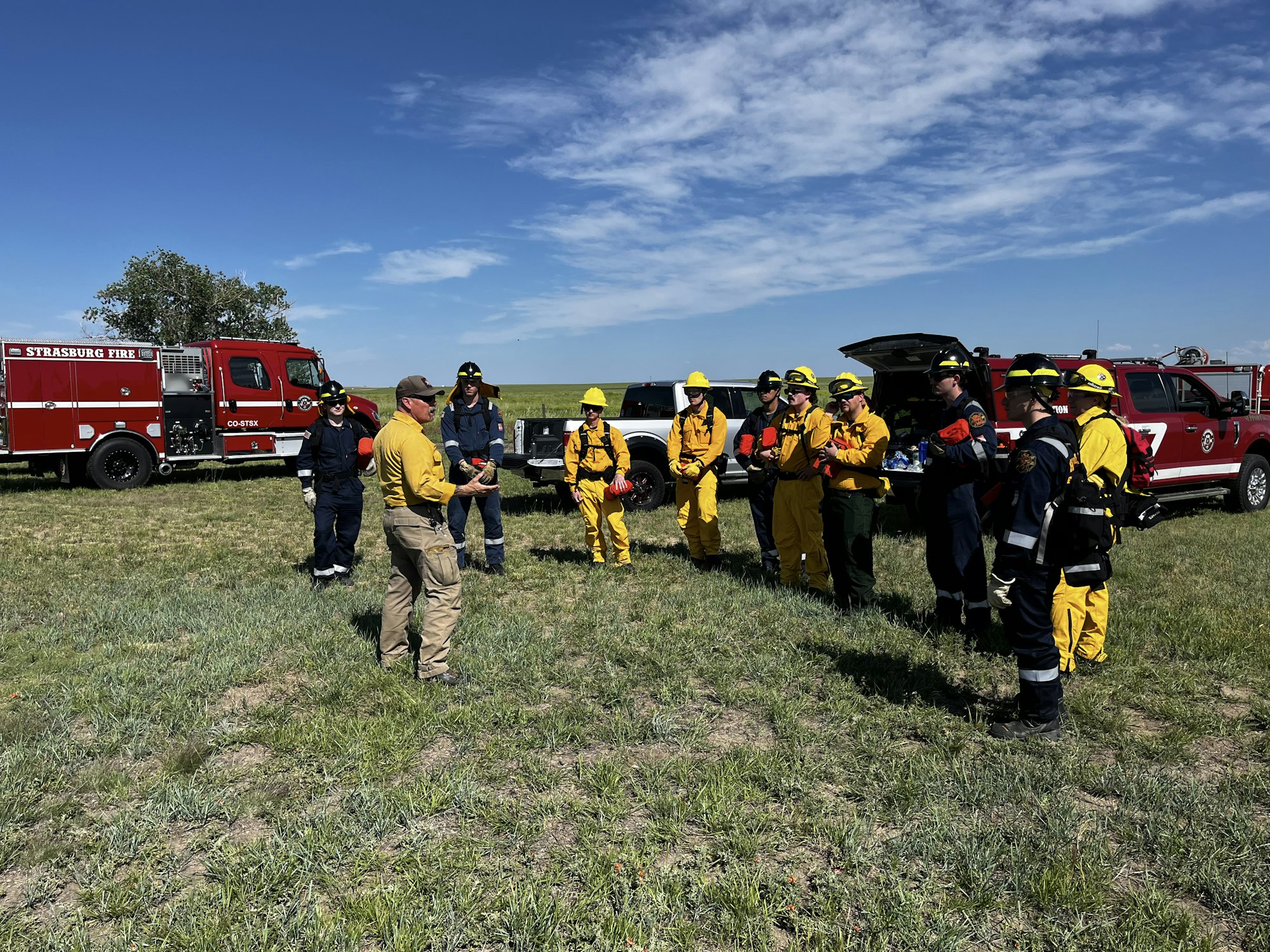 Firefighters in gear gather outdoors near red fire trucks under a blue sky.