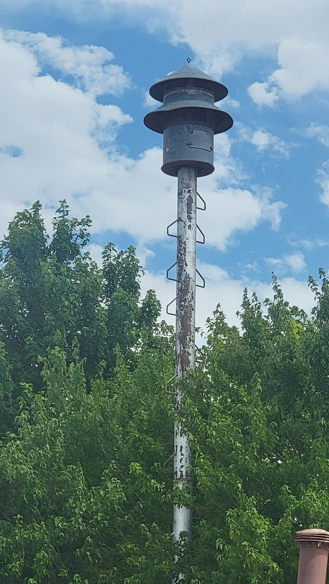 A tall metal pole with a siren at the top, surrounded by trees and a blue sky with clouds.
