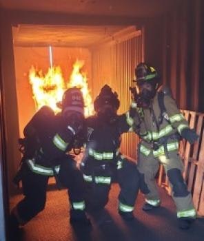 Three firefighters in gear inside a building with visible flames behind them.