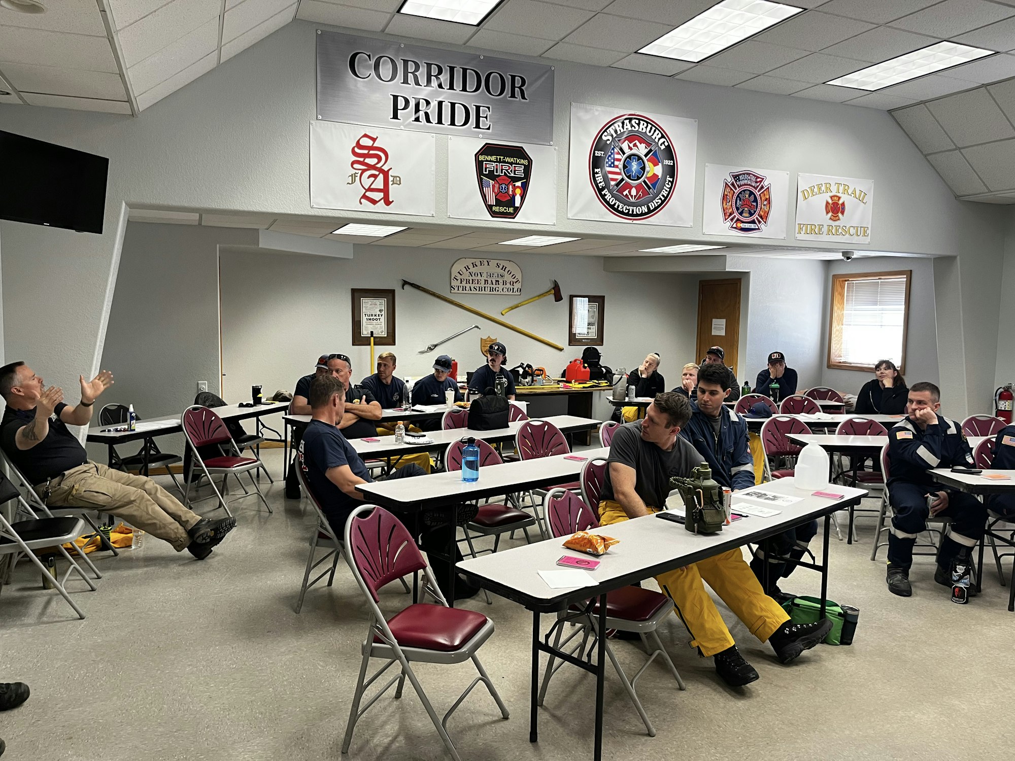 A classroom with firefighters seated at tables, listening to a speaker. Banners for various fire departments are on the wall.