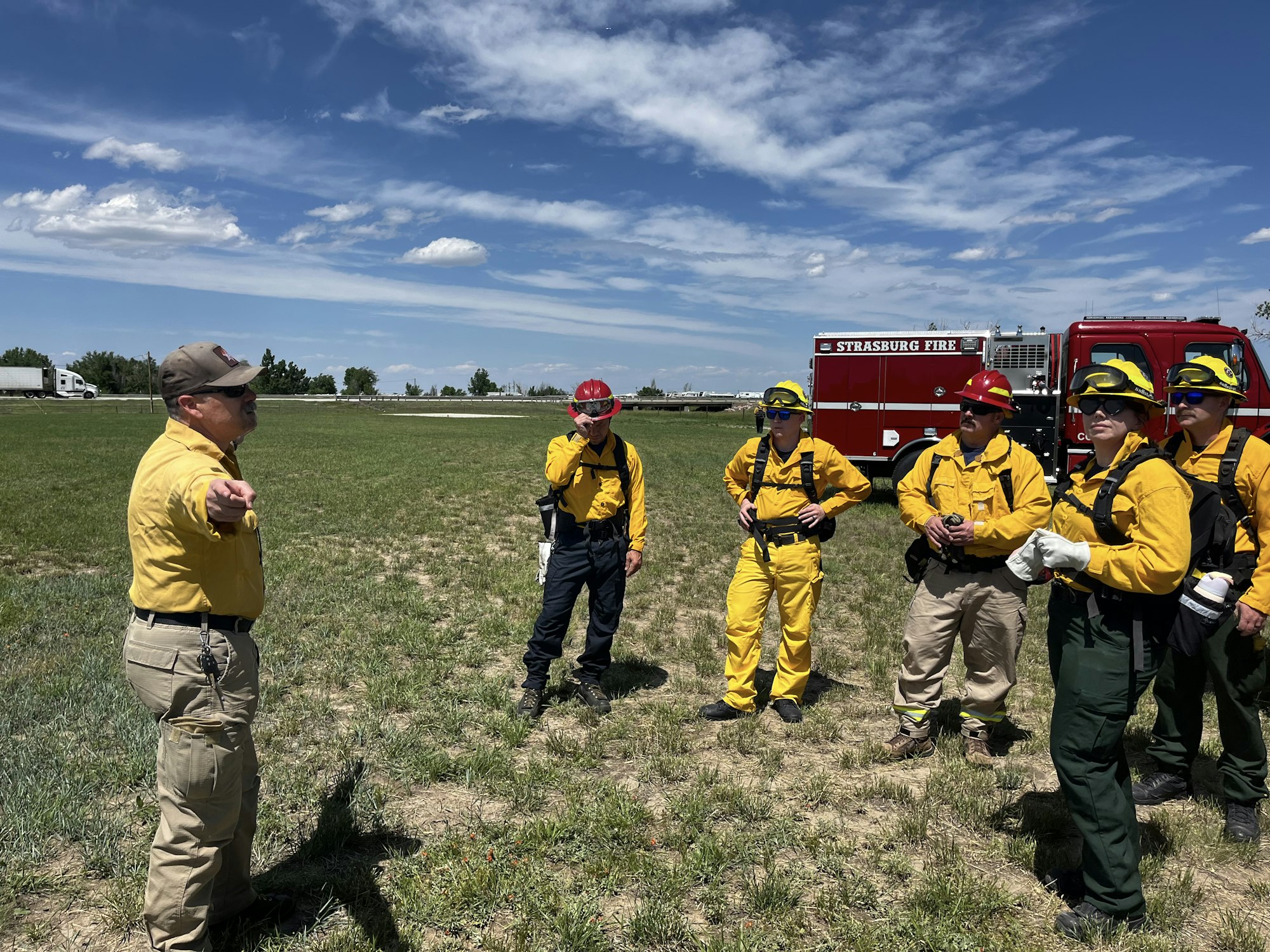 A group of firefighters in yellow and red gear stands in a grassy field with a fire truck in the background.