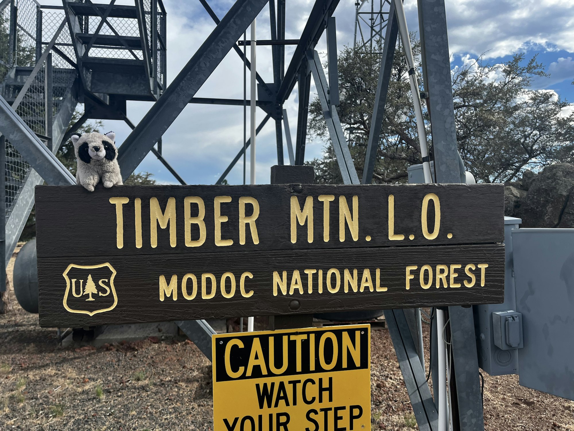Sign for Timber Mountain Lookout in Modoc National Forest with a toy raccoon on it and a caution sign below.