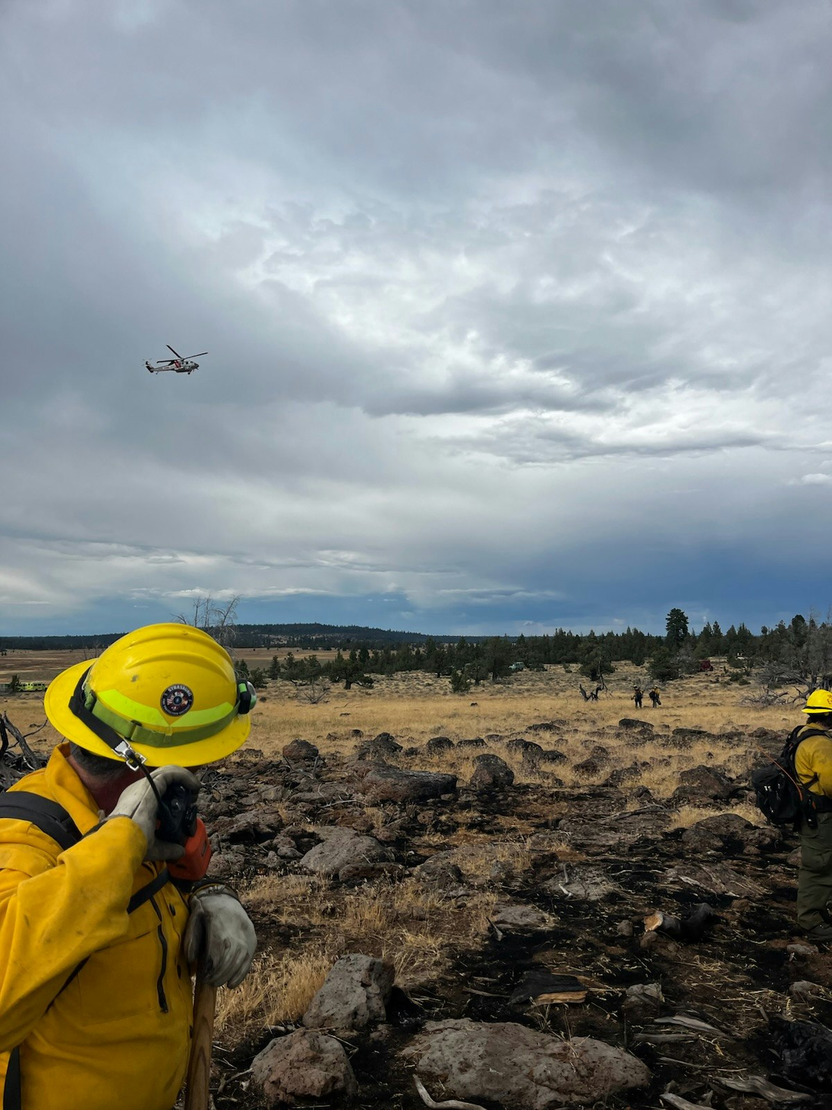 Firefighters in yellow gear in a rocky field, with a helicopter flying overhead under a cloudy sky.